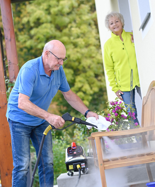 Ein älterer Mann reinigt die Holzbank vor seinem Haus mit Thermostar-Trockendampf, während seine Frau zusieht.