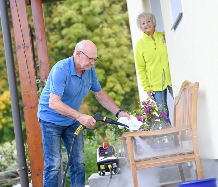 Ein älterer Mann reinigt die Holzbank vor seinem Haus mit Thermostar-Trockendampf, während seine Frau zusieht.